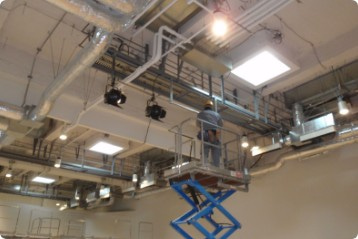 舞台照明設備の天井部分近くで黄色いヘルメットとグレーの作業着で作業をする一人の男性の写真