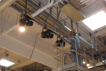 舞台照明設備の天井にある照明器具の近くで黄色いヘルメットとグレーの作業着で作業をする一人の男性の写真
