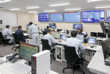 5～6人のグレーの服を着た男性がPCの置かれた席に座り、操作しながら各種モニターをチェックするなどの監視作業を行っている様子の写真