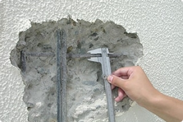 コンクリートの壁の一部がえぐれており、その部分にノギスを当てて測定している写真