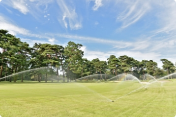 背景に青空と木々の緑が綺麗なゴルフ場のコースに散水設備により水がまかれている様子を撮影した写真