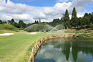 背景に青空と木々の緑、そして池もあるゴルフ場のコースに散水設備により水がまかれている様子を撮影した写真