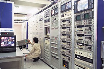 左右の壁いっぱいに設置された受信点装置・ヘッドエンド装置の中で作業している一人の男性の写真